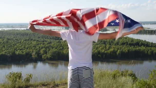 Blonďatý chlapec mává národní vlajkou USA venku nad modrou oblohou na břehu řeky - Záběry, video