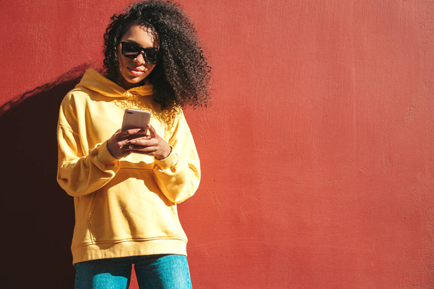 Beautiful black woman with afro curls hairstyle.Smiling model in yellow hoodie. Sexy carefree female posing on the street near red wall in sunglasses. Looking at smartphone screen, using apps - Foto, imagen