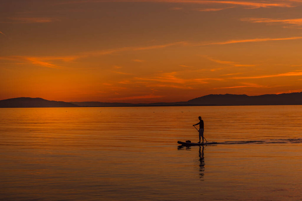 Persona no reconocida disfrutando de la puesta de sol en la costa desde una mesa de body board. - Foto, Imagen