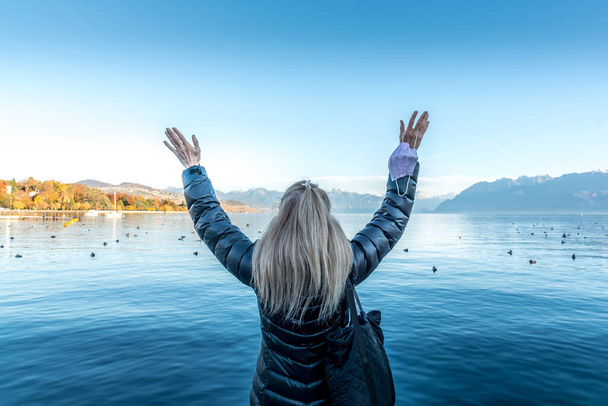 Faceless blonde woman in a lake raising her arms and holding her face mask. - Photo, Image