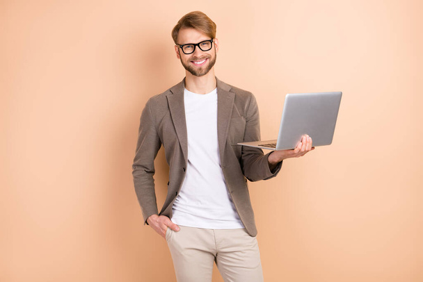 Photo of optimistic nice blond hair man hold laptop wear spectacles grey jacket isolated on beige color background - 写真・画像