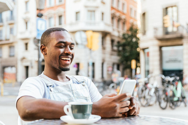 Happy Afro-Amerikaanse jongeman met behulp van zijn smartphone op het terras van een bar - Foto, afbeelding
