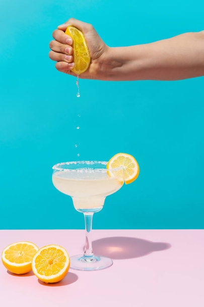 mão de uma mulher desconhecida espremendo um limão em um coquetel margarita em um fundo azul e rosa. conceito de bebida, México e verão. - Foto, Imagem