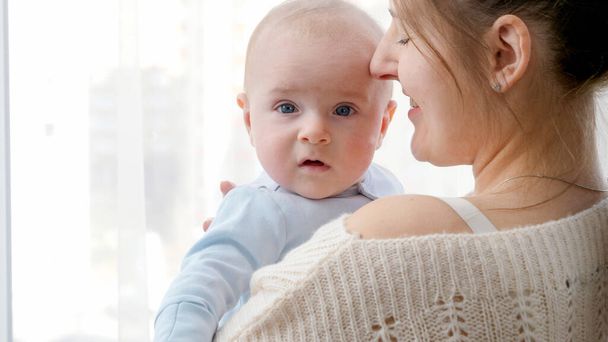 Closeup portrait of baby and mother face hugging and embracing at big window at home. Concept of family happiness and child development - 写真・画像