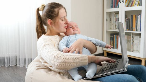 幼い母親はノートパソコンで自宅のオフィスで働きながら赤ちゃんを撫でていた。家族の幸福と子供の発達の概念. - 写真・画像
