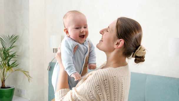 Happy laughing and smiling baby boy looking at mother while she is holding and shaking him. Concept of family happiness and child development. - 写真・画像