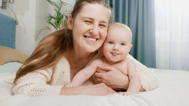 幸せな母親と一緒にベッドに横たわって、カメラを見て笑顔の小さな赤ちゃんの男の子。子育て・育児・家族の幸せの概念 - 写真・画像