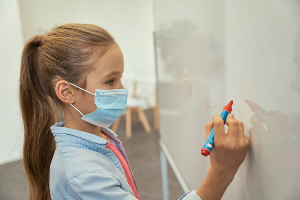 Portrait of little schoolgirl wearing protective mask during coronavirus pandemic writing numbers on board in elementary school classroom - Fotó, kép