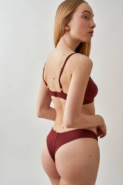 Back view of attractive young slim caucasian woman with beautiful perfect body wearing burgundy underwear looking aside, posing isolated over light gray background - 写真・画像