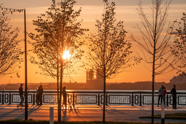 Silhouettes of people walking along the embankment against the background of the setting sun and glare on the water. Sunset on the city promenade. - Photo, image