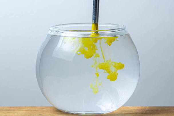 кисть с желтой краской погружается в банку с водой на белом фоне. краска распространяется в воде. - Фото, изображение