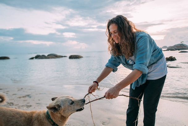 Meisje heeft plezier spelen met een hond op het strand bij zonsondergang. Een jonge Europese vrouw loopt met een hond op de achtergrond van de zee en de ondergaande zon. - Foto, afbeelding