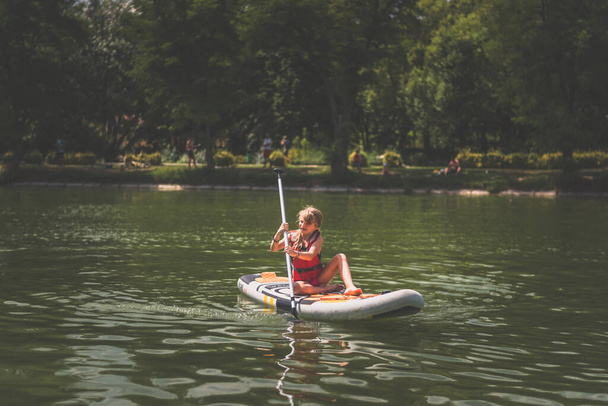 young girl in red safety inflatable vest paddling on paddle board on water in hot sunny summer day - Photo, image