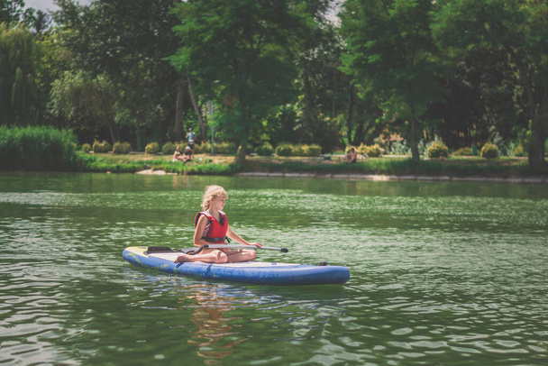menina nova no colete inflável da segurança vermelha que rema na placa da pá na superfície da água no dia ensolarado quente do verão na natureza verde ao ar livre - Foto, Imagem