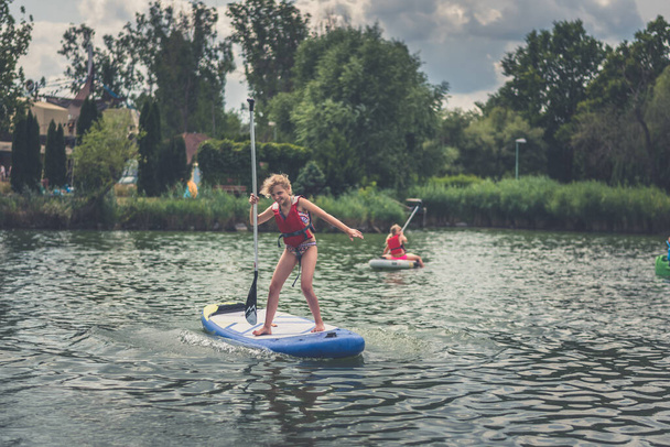 young girl in red safety inflatable vest paddling on paddle board on water surface in hot sunny summer day in green nature outdoors - Photo, Image