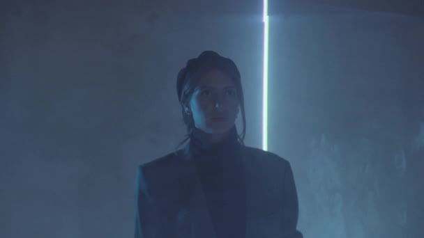Young beautiful woman in trendy outfit standing in dark studio with moving laser light projected on her and looking away pensively - Materiał filmowy, wideo