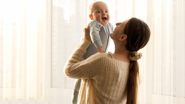 Bonne mère souriante levant, jouant et embrassant son bébé contre une grande fenêtre à la maison. Concept de bonheur familial et de parentalité - Photo, image