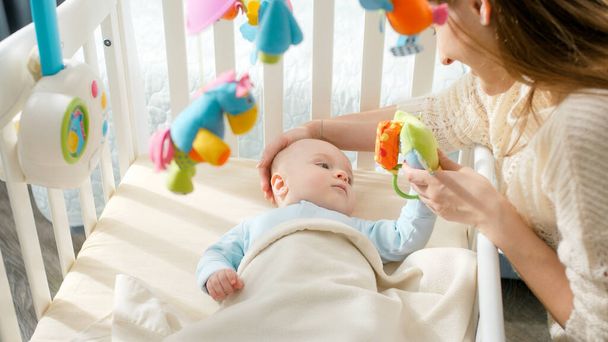 Young mother playing toys and stroking her little baby son lying in cradle. Concept of parenting, family happiness and baby development - 写真・画像