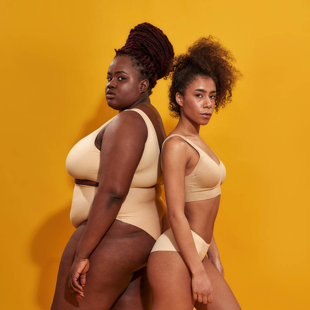 Two strong african american women in beige underwear with different body size standing together back to back, posing isolated over yellow background - Фото, зображення