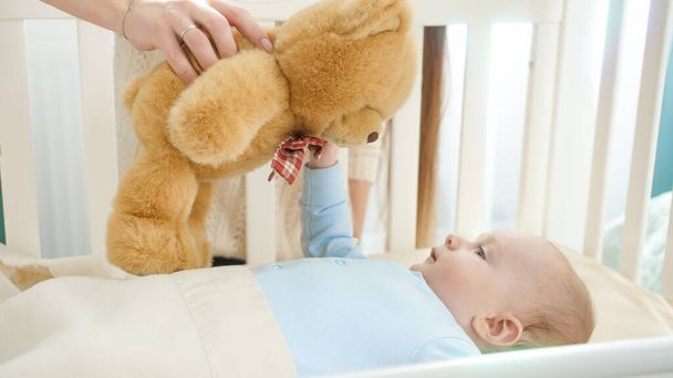 Netter kleiner Junge, der auf Spielzeug-Teddybär schaut, der von Mutter gehalten wird. Konzept von Elternschaft, Familienglück und Babyentwicklung - Foto, Bild