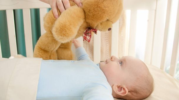 Young mother playing with teddy bear and her little baby boy lying in crib. Concept of parenting, family happiness and baby development - Photo, image