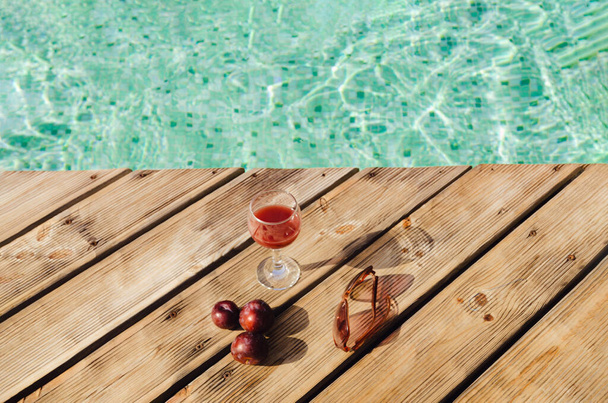 A glass with fruit juice, sun glasses and some plums on wood boards along the edge of a pool with blue mosaic tiles. Summer vibes and relaxation minimal composition. - Foto, Imagem