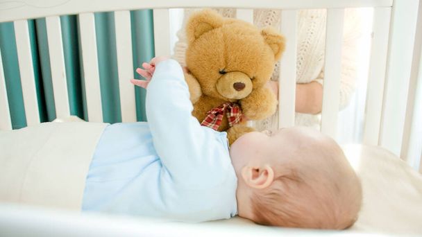 Toy teddy bear playing with little baby boy lying in cradle. Concept of parenting, family happiness and baby development - Foto, imagen