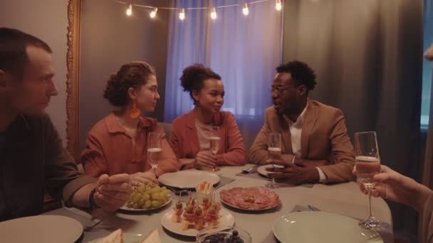 Zoom-out slow mo de grupo de jóvenes amigos multiétnicos sentados en la cena festiva tostadas con copas de champán celebrando ocasión especial - Imágenes, Vídeo