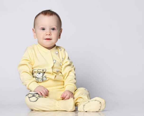 Μικρό χαριτωμένο μωρό κάθεται στο πάτωμα στο στούντιο σε ένα καλοκαιρινό βαμβακερό κοστούμι, σε ένα βαμβακερό κοστούμι σε ένα ελαφρύ φόντο. απομονωμένα - Φωτογραφία, εικόνα