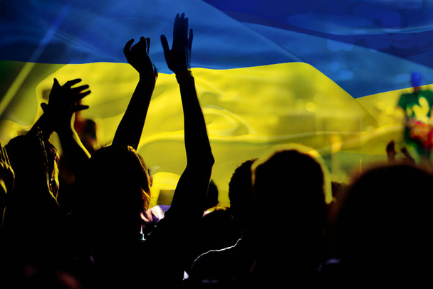 football fans supporting Ukraine - crowd celebrating in stadium with raised hands against Ukraine flag - Photo, Image