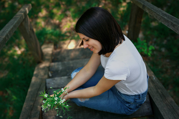 Uma mulher bonita nova sentou-se para descansar em uma ponte de madeira velha na floresta e olha para seu buquê coletado de flores selvagens. - Foto, Imagem
