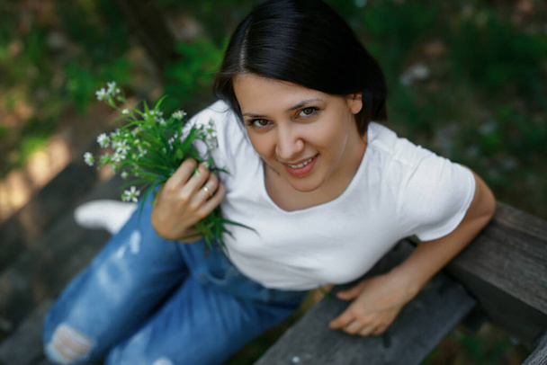 Молода жінка в рваних блакитних джинсах з букетом білих диких квітів сидить на старому дерев'яному пішохідному мосту
. - Фото, зображення