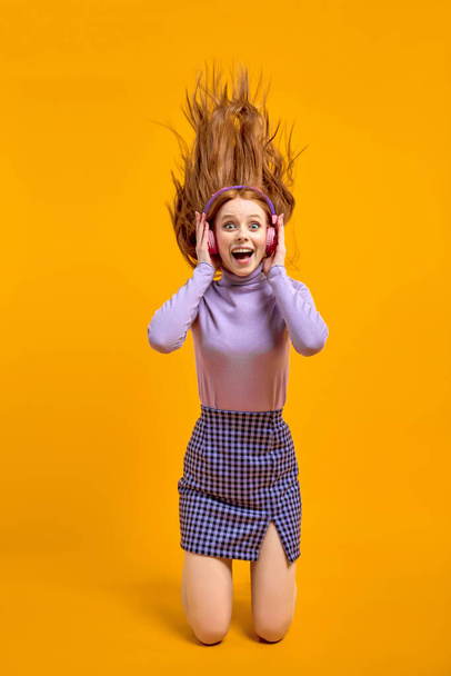 Emotional young excited female in headphones with flying hair isolated on yellow - Photo, Image