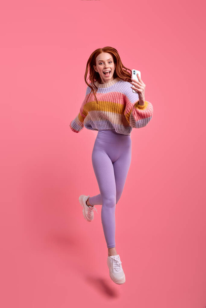 caucasian Shocked redhead woman in casual clothes jumping and taking photo on smartphone - Foto, afbeelding
