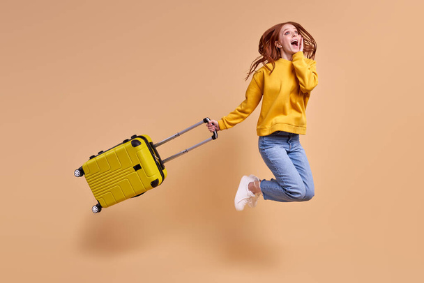 Excited beautiful redhead woman is going to have vacation jumping with cabin bag - Photo, image