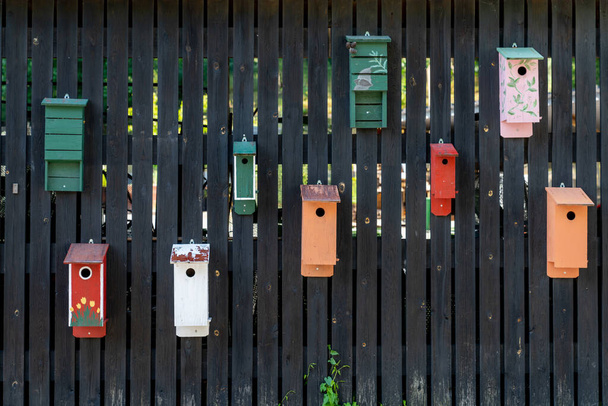 A dark wooden fence with many colorful bird houses mounted on it - Fotó, kép