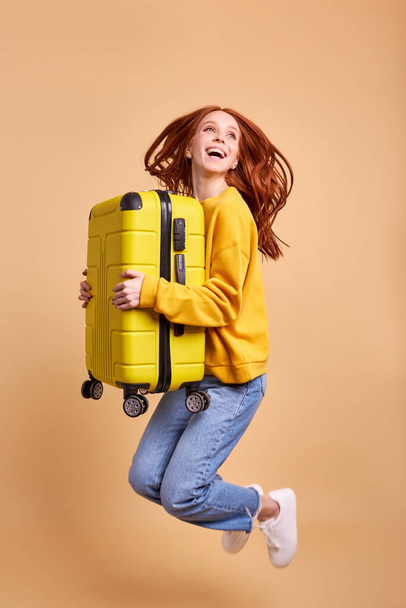 Young tourist redhead woman with suitcase, jumping in casual outfit isolated in studio - Valokuva, kuva