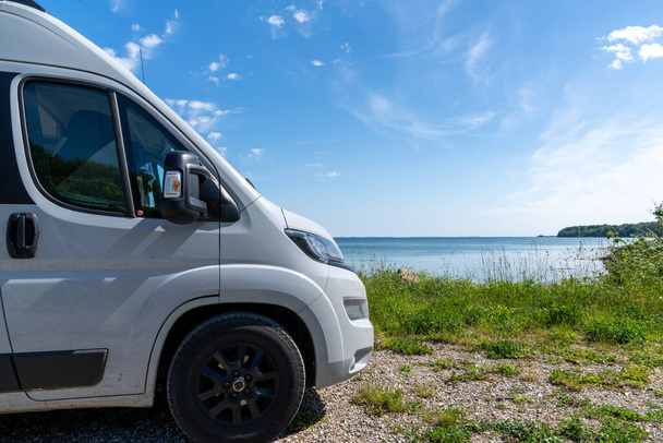 The front end of a gray camper van parked next to a picturesque shore with turquoise water and forest under a blue sky - Photo, Image