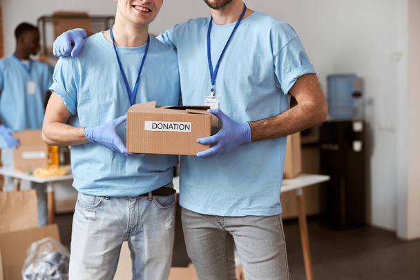Foto recortada de dos chicos caucásicos, voluntarios masculinos en uniforme azul, guantes protectores posando juntos y sosteniendo la caja de cartón para Donación - Foto, Imagen