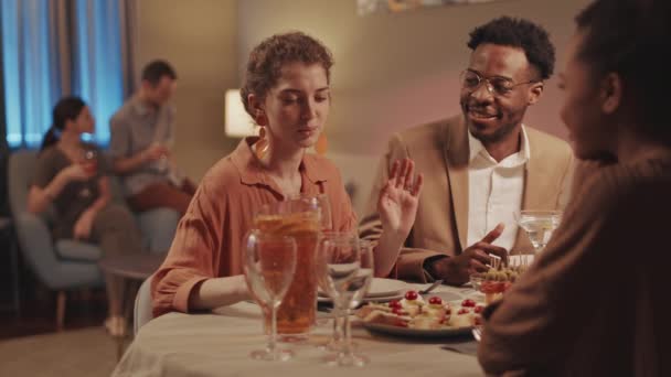 Střední zpomalení mladého Afroameričana muže a dvě krásné ženy sedí u jídelního stolu komunikovat, zatímco pár jejich přátel chatuje v rozmazaném pozadí během domácího večírku - Záběry, video