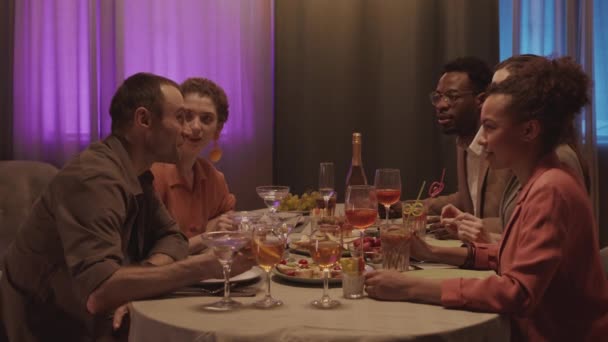 Mittlere Slow-Motion von fünf fröhlichen multiethnischen Freunden, die um einen schönen festlichen Tisch sitzen und miteinander gute Zeit in einer gemütlichen Wohnung mit Neonbeleuchtung an Vorhängen verbringen - Filmmaterial, Video