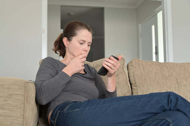 Σοκαρισμένη ενήλικη γυναίκα (ηλικίας 30-40 ετών) που διαβάζει κείμενο μασάζ στο κινητό τηλέφωνο στο σαλόνι του σπιτιού. - Φωτογραφία, εικόνα