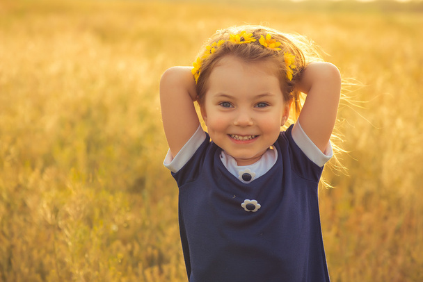The beautiful little girl in a blue dress against a field laughs, holds hands behind the head - Фото, зображення