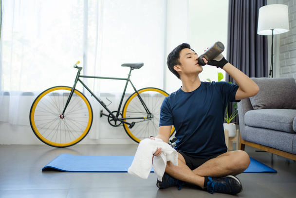 Asiatischer Mann trinkt Wasser nach dem Sport im heimischen Wohnzimmer. Er fühlte sich müde. - Foto, Bild