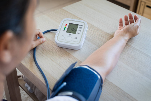 アジアの若い女性は自宅で血圧計で血圧と心拍数を自分でチェックしています。健康と医療の概念. - 写真・画像