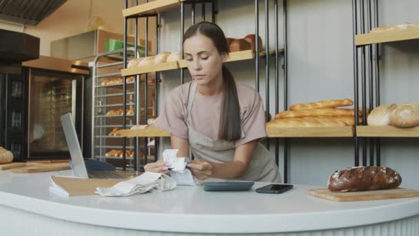 Slowmo sledování záběr mladé ženské pokladní při pohledu na účtenky a pomocí kalkulačky v útulné pekárně - Záběry, video