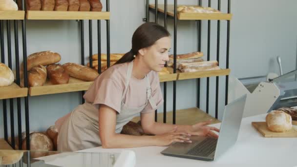 Slowmo śledzenie ujęcia młodej sklepikarki w fartuchu stojącej za ladą w przytulnej piekarni i przy użyciu laptopa - Materiał filmowy, wideo