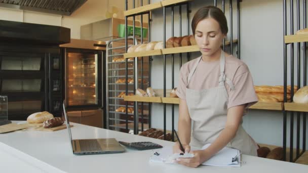 Slowmo střední záběr mladé ženy v zástěře stojící za pultem v útulné pekárně a kontrolu stvrzenek - Záběry, video
