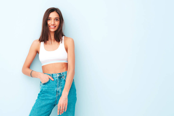 Beautiful smiling woman dressed in white jersey top shirt and jeans. Sexy carefree cheerful model enjoying her morning. Adorable and positive female posing near light blue wall in studio - Foto, imagen