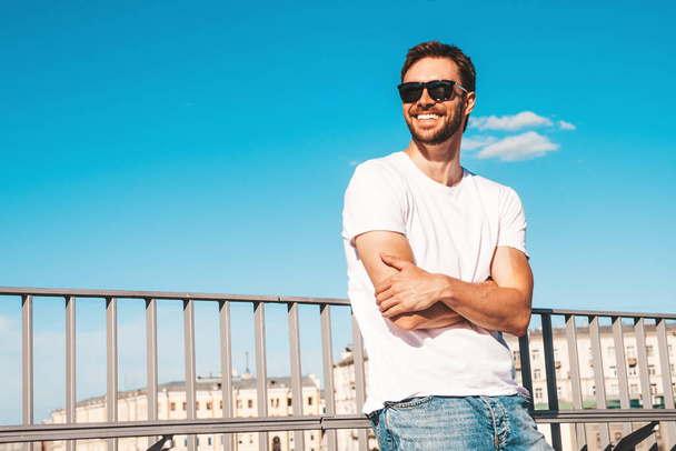 Portrait of handsome smiling  hipster lambersexual model.Stylish man dressed in white T-shirt. Fashion male posing behind blue sky on the street background in sunglasses.Crossed arms - Zdjęcie, obraz